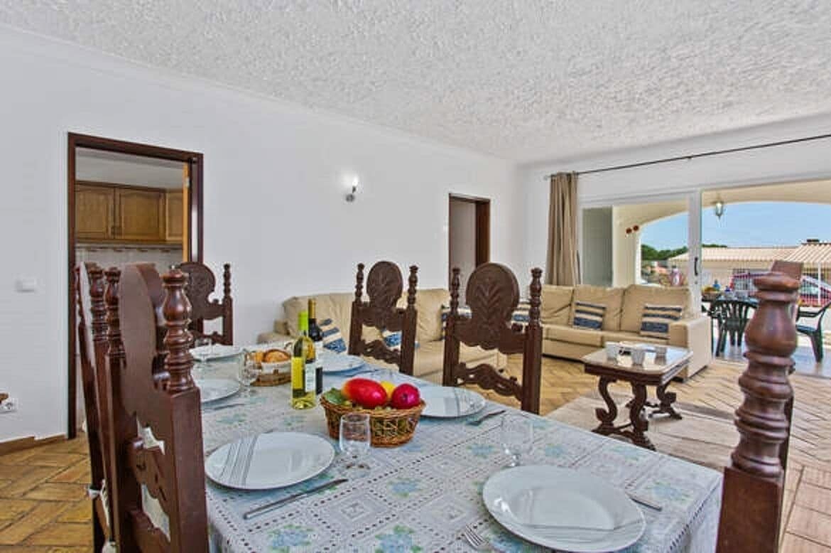 Comedor con una mesa dispuesta para cuatro personas, rodeada de sillas de madera. En la mesa hay una cesta de fruta y una botella de vino. La habitación tiene un ventanal con vistas al exterior.