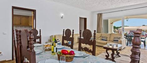 Comedor con una mesa dispuesta para cuatro personas, rodeada de sillas de madera. En la mesa hay una cesta de fruta y una botella de vino. La habitación tiene un ventanal con vistas al exterior.