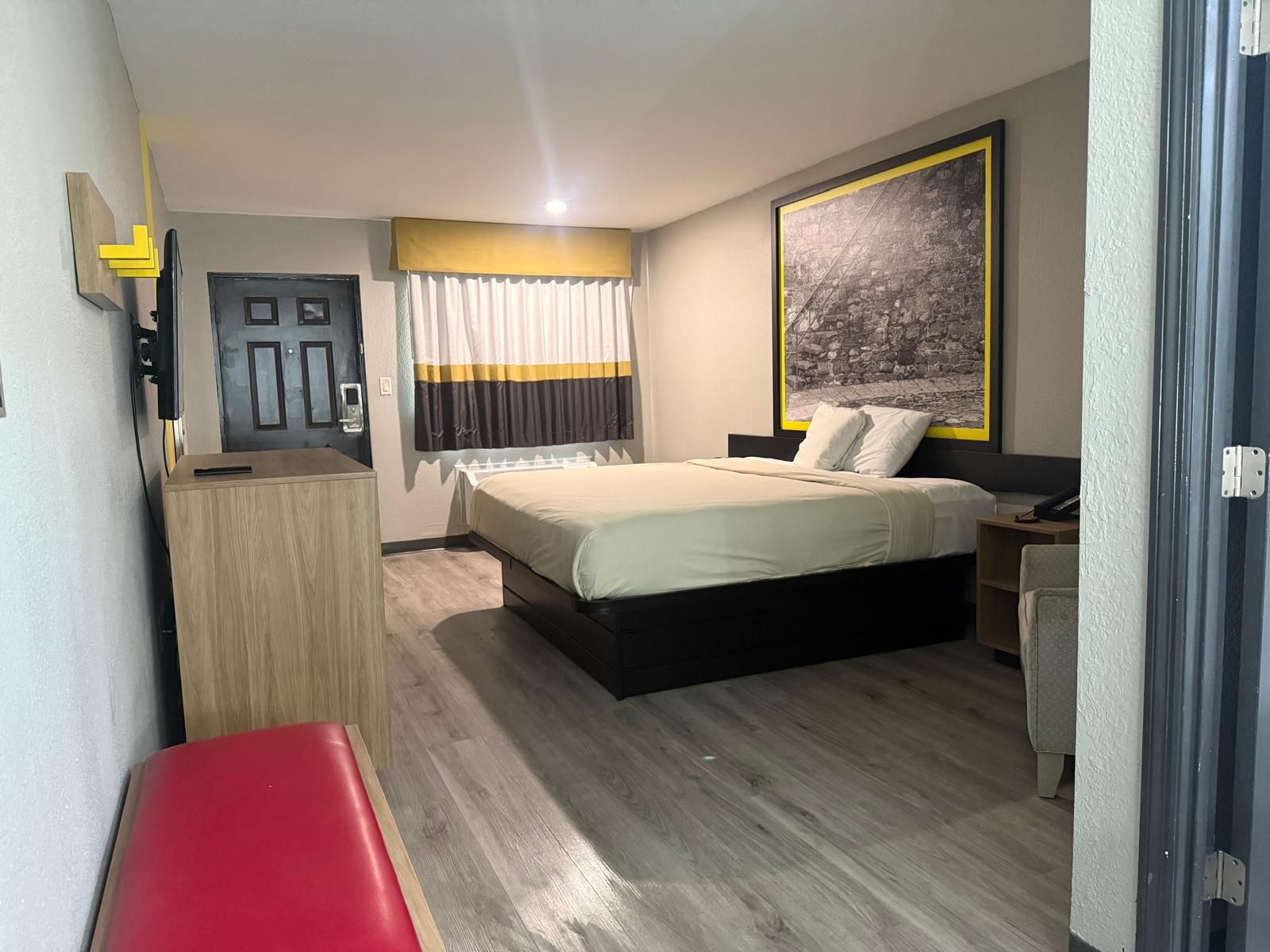 Habitació d'hotel amb llit, escriptori, cadira, banqueta, porta i finestra amb cortines.
