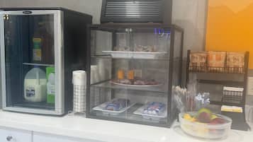 A self-service breakfast station with a refrigerator, a display case with food items, and a bowl of fruit.