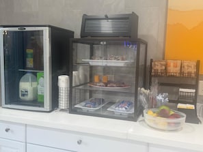 Un espace de petit-déjeuner en libre-service comprenant un réfrigérateur, un présentoir avec des produits alimentaires et un saladier de fruits.
