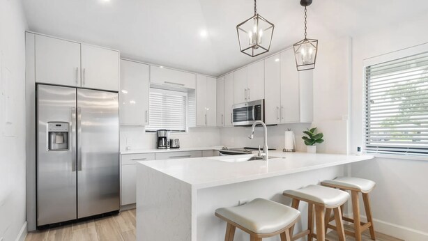 A modern kitchen with white cabinetry, stainless steel appliances, a central island with stools, and pendant lighting. - Private Cottage at PGA National (Palm Beach Gardens)
