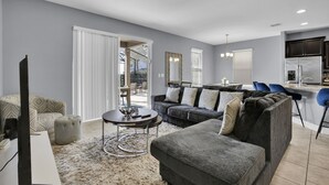 A modern living room with a grey sofa, ottoman, and a coffee table. There is a kitchen area with dark cabinets and a refrigerator. - ✨ Luxurious 6BR Villa with Private Pool & Game Room near Disney 🏊‍♂️🎮 (Davenport)