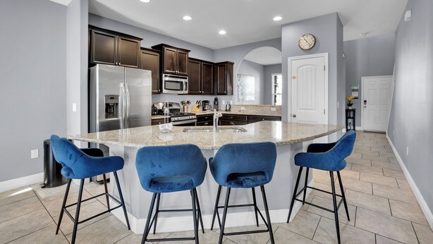 A modern kitchen with a central island, bar stools, stainless steel appliances, and a clock on the wall. - ✨ Luxurious 6BR Villa with Private Pool & Game Room near Disney 🏊‍♂️🎮 (Davenport)
