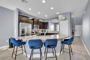 A modern kitchen with a central island, bar stools, stainless steel appliances, and a clock on the wall. - ✨ Luxurious 6BR Villa with Private Pool & Game Room near Disney 🏊‍♂️🎮 (Davenport)