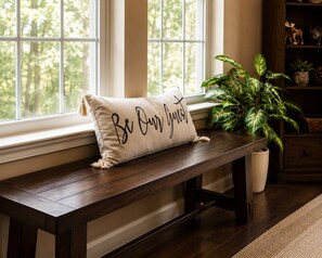 Een bank met een kussen waarop 'Be Our Guest' staat, in een kamer met een groot raam, een potplant en een houten kast.