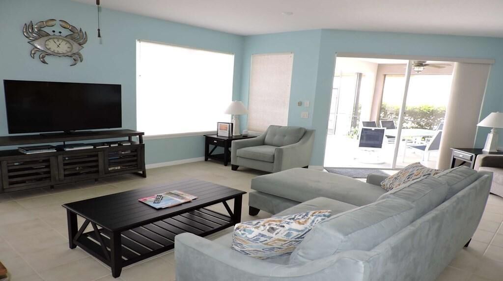 A living room with a TV, a sofa, a coffee table, and a chair. There is a window and a door leading to an outdoor area.