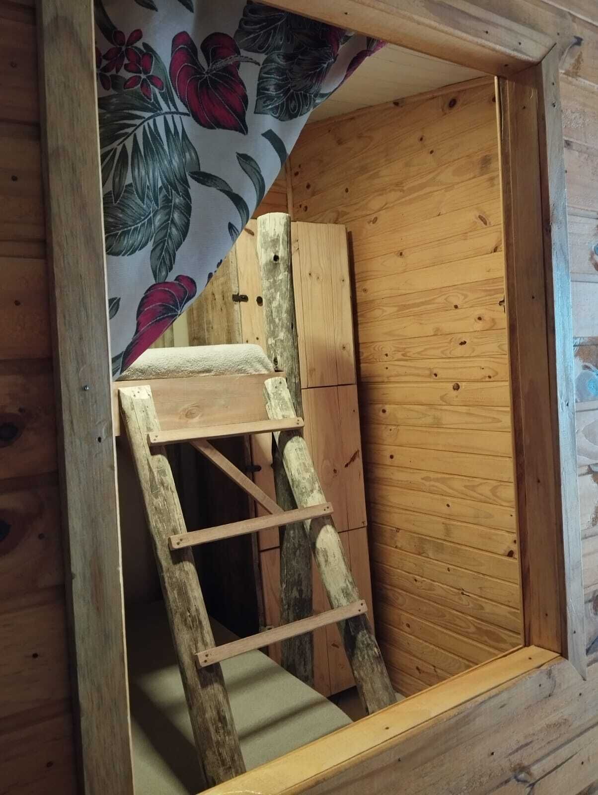 A wooden ladder leading to a small wooden platform inside a cabin with a floral-patterned fabric hanging above.