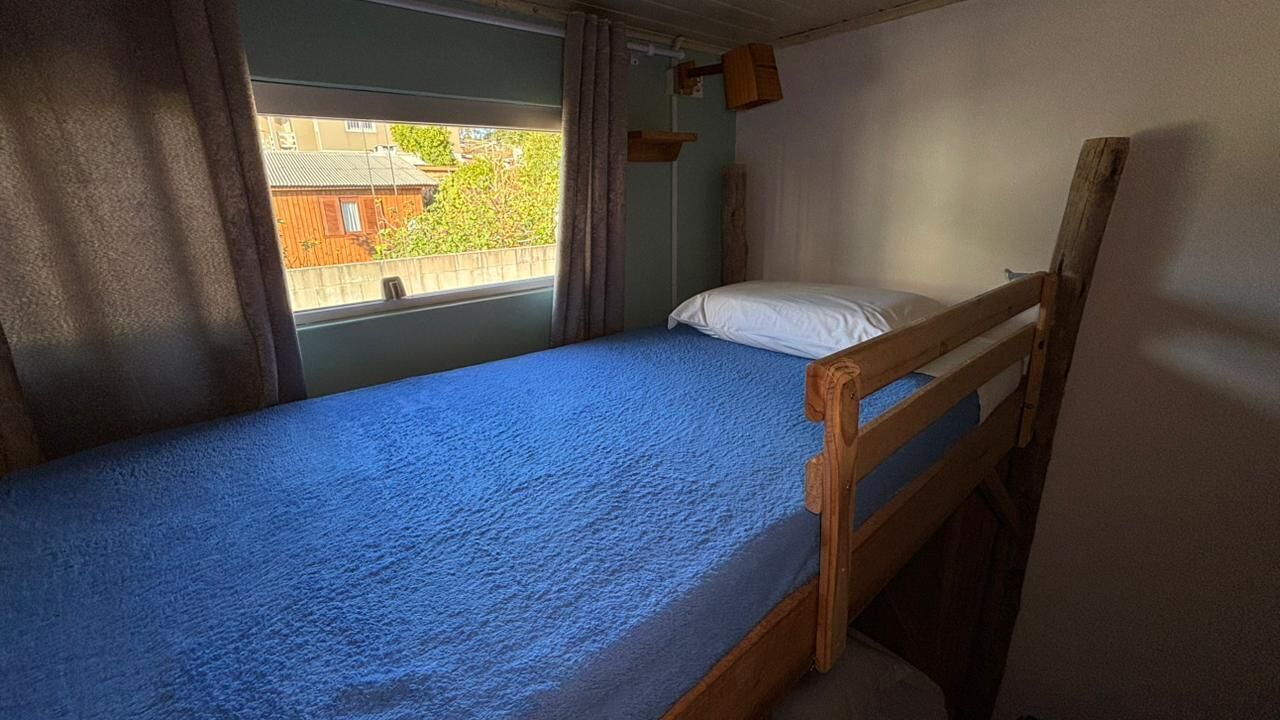 A single bed with a blue mattress, a wooden headboard, a window with curtains, and a view of a building outside.