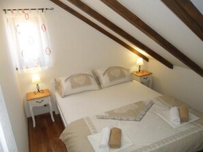 Quarto com cama, duas mesas de cabeceira, um criado-mudo, janela com cortinas e uma viga de madeira no teto.