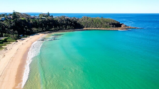 Un paysage côtier avec une plage de sable, des eaux turquoise et limpides, et une colline boisée en arrière-plan.