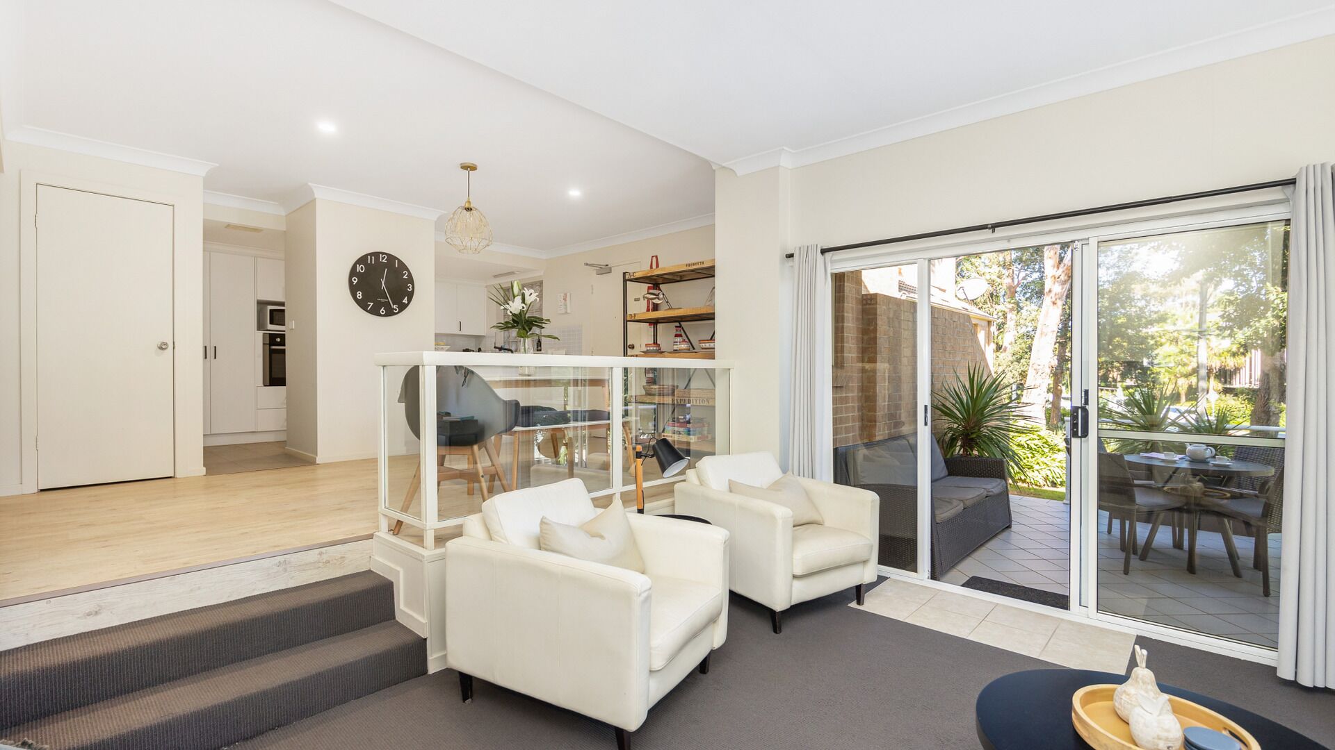 A modern living room with white sofas, a glass table, and a kitchen in the background.