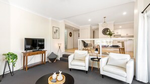 Un salon moderne doté d’un téléviseur à écran plat, de deux fauteuils blancs, d’une table basse et d’un coin repas séparé par une cloison en verre.