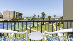 A balcony with two chairs and a small table, overlooking a tennis court and a beach.