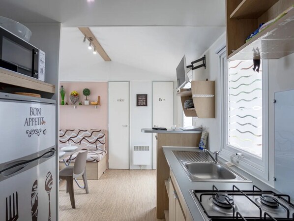 A compact kitchen with a refrigerator, stove, and microwave. There is a dining area with a chair and a small table. The kitchen has a window with blinds and a door labeled "Bain".