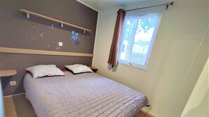 A bedroom with a bed, a nightstand, a wall-mounted shelf, and a window with curtains. - Mobile home near the beaches (Saint-Jean-de-Monts)
