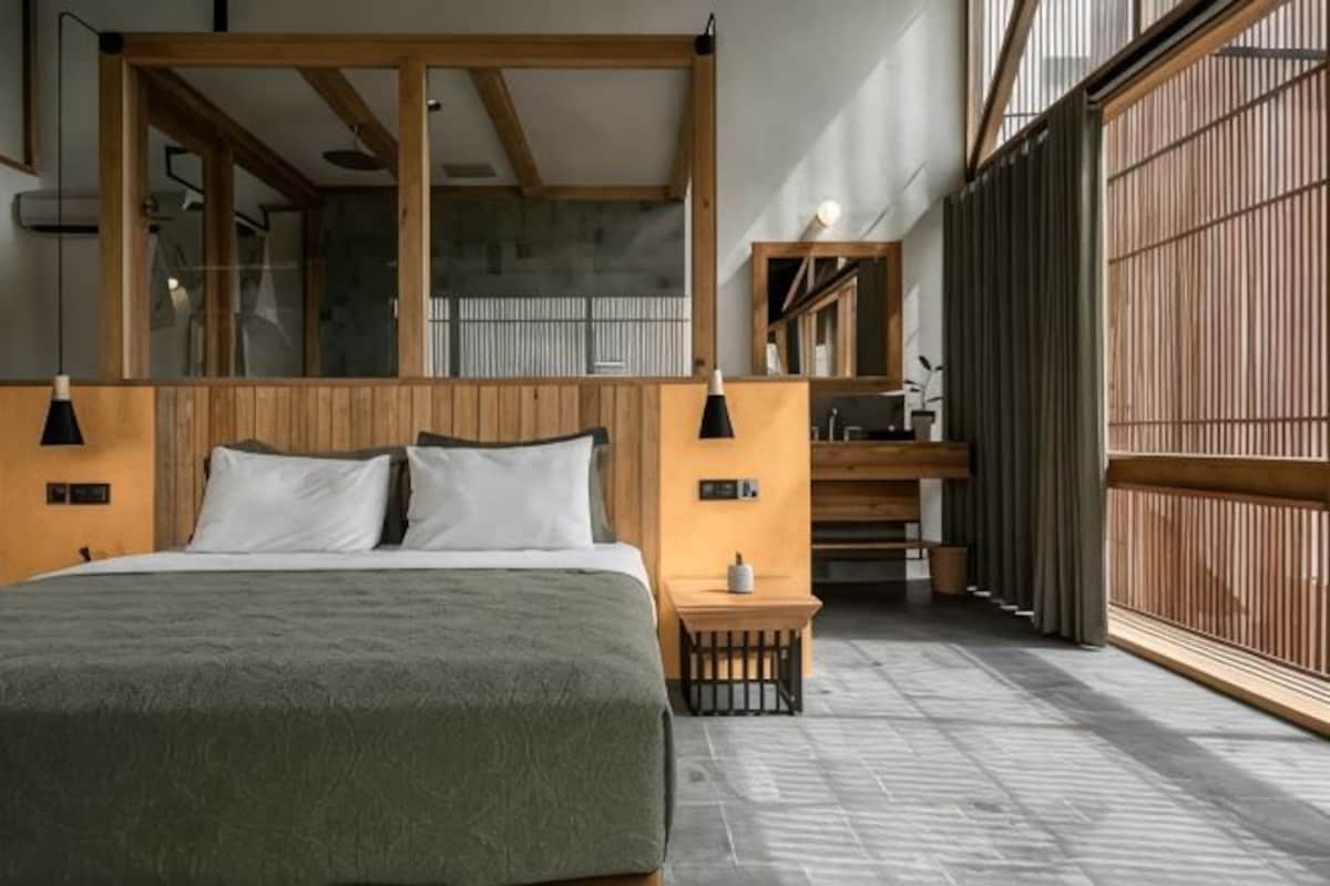 Une chambre à coucher avec un grand lit, une tête de lit en bois, des tables de chevet, une petite table de nuit et une vue sur l’extérieur, accessible par une porte coulissante.