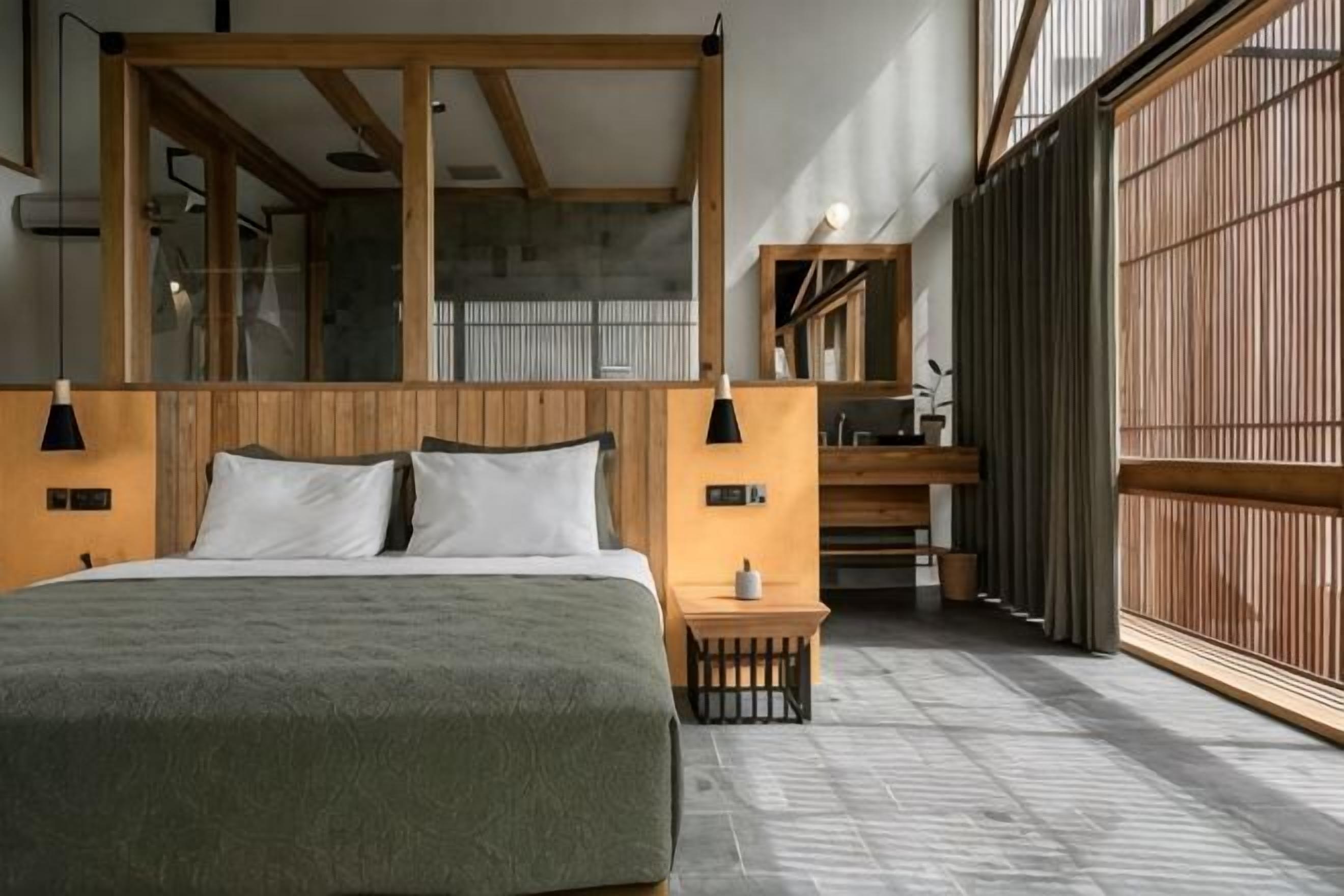 Une chambre à coucher avec un grand lit, une tête de lit en bois, des tables de chevet, une petite table de nuit et une vue sur l’extérieur, accessible par une porte coulissante.