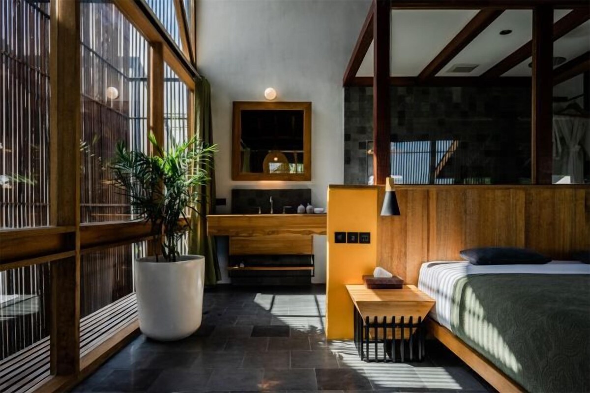A room with a bed, a wooden bench, a potted plant, and a wall-mounted mirror.