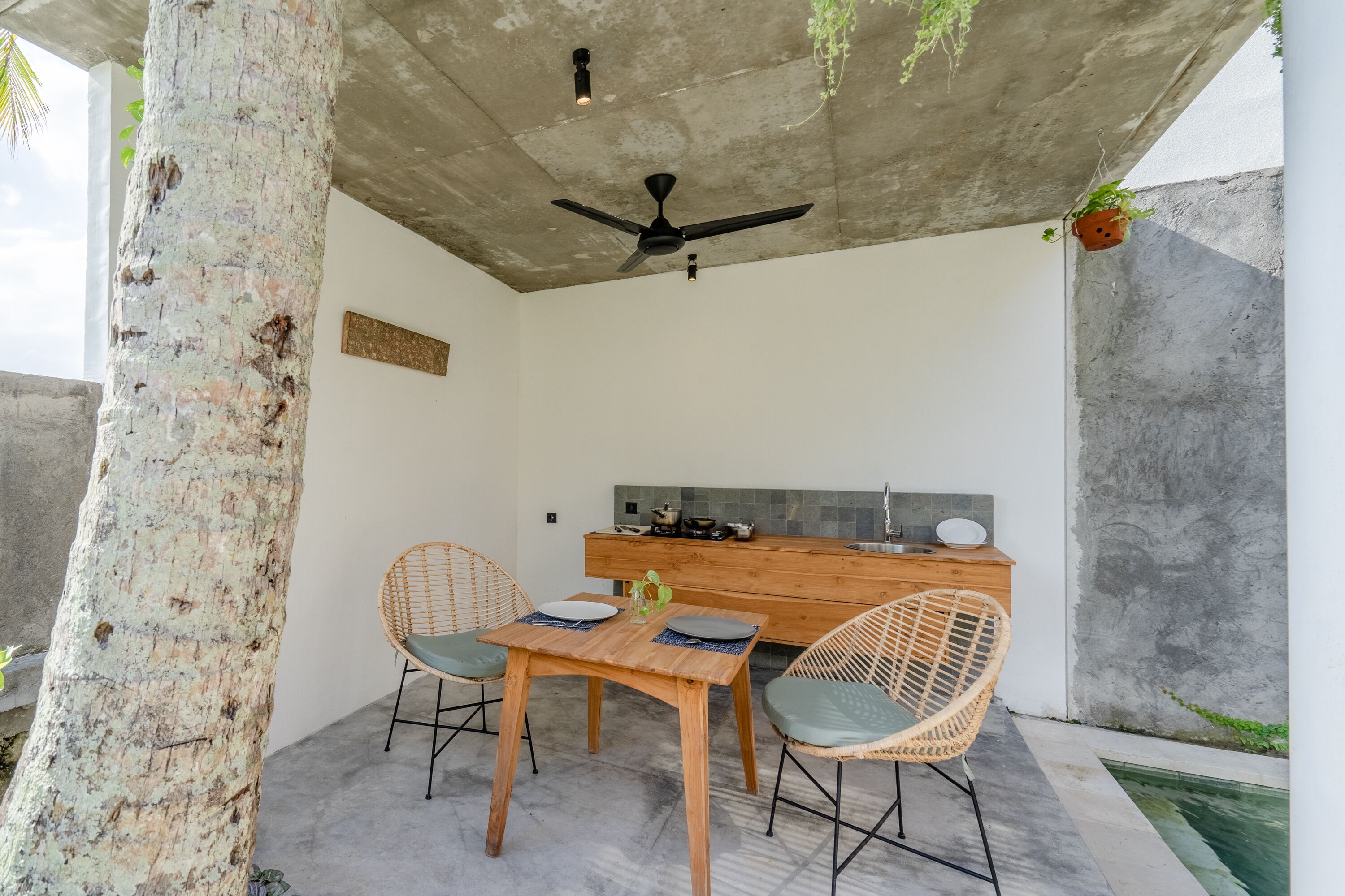 A modern outdoor dining area with a wooden table and wicker chairs, a ceiling fan, and a kitchen counter in the background.