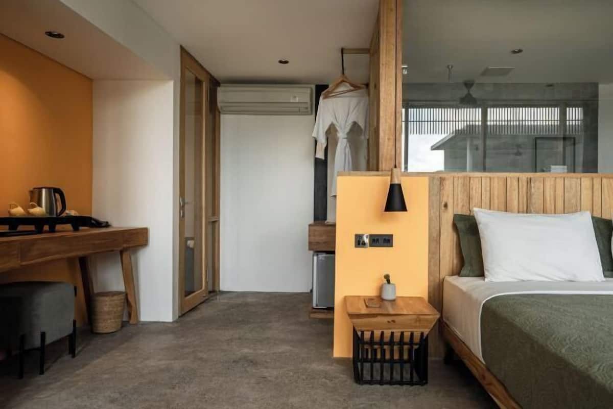 A modern hotel room with a wooden desk, a bed with green bedding, a hanging robe, and a small table with a lamp.