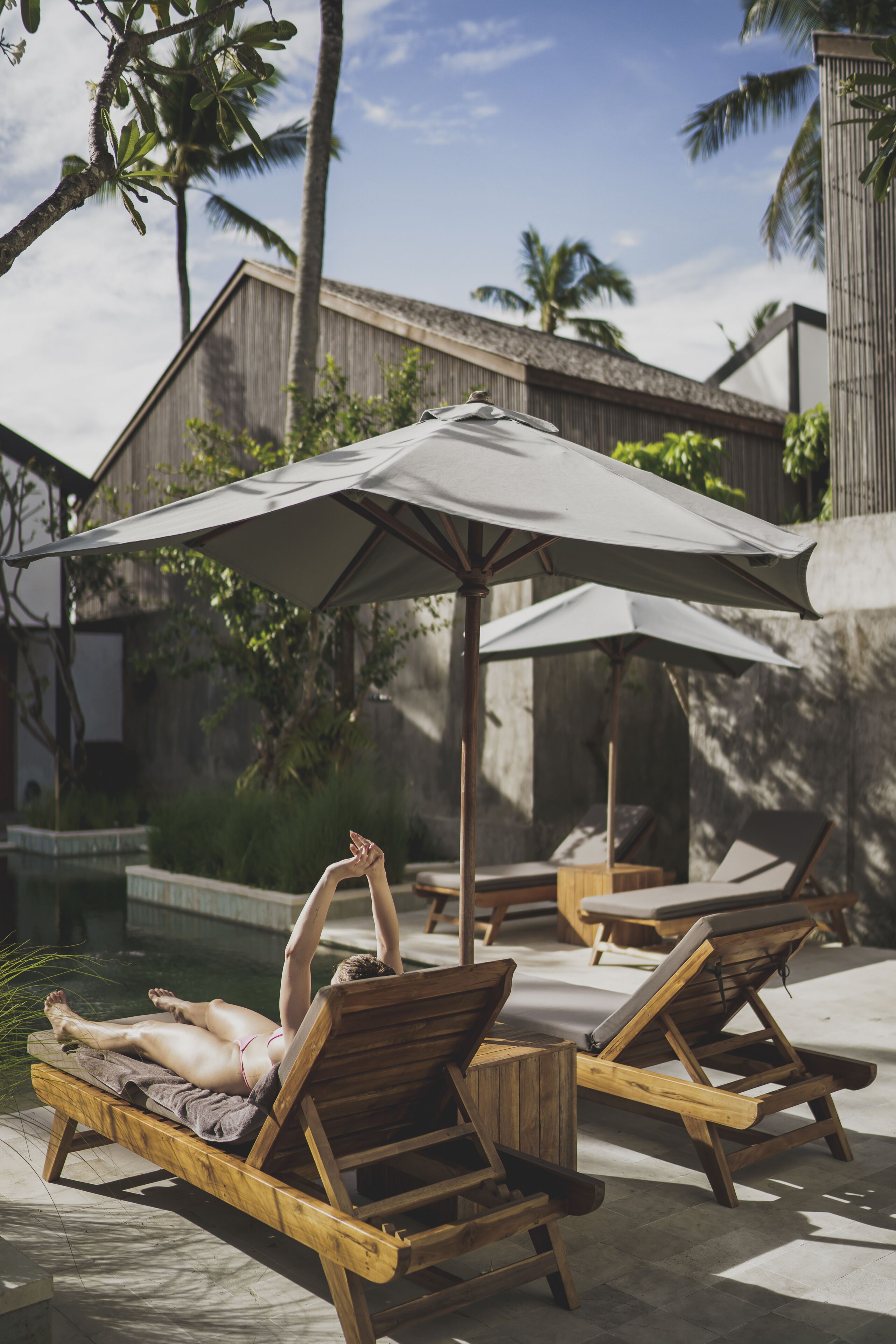 Un espace aménagé au bord de la piscine, avec des chaises longues en bois et des parasols, entouré de palmiers et d’un bâtiment.