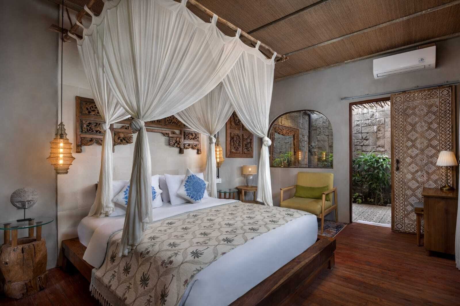 A bedroom with a four-poster bed, a canopy, a wooden floor, a small table with a lamp, and a chair.