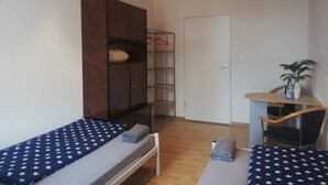 Una habitación pequeña y funcional con dos camas individuales, un armario, una silla y una mesita con una planta.