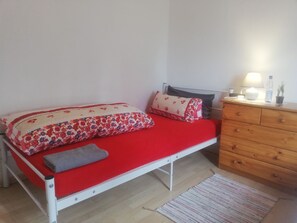 Habitación con cama individual, colcha roja, cómoda de madera, mesita de noche con lámpara y una alfombra pequeña.