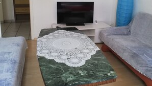 Una sala de estar con una televisión sobre un mueble bajo, una mesa de centro con un posavasos de encaje, dos sofás grises y un jarrón azul.