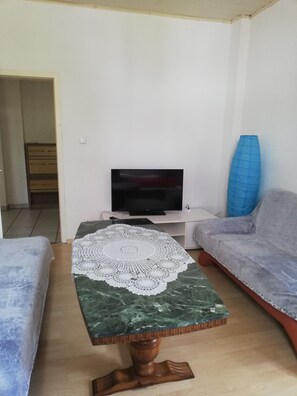 A living room with a television on a stand, a coffee table with a lace doily, two grey sofas, and a blue vase.