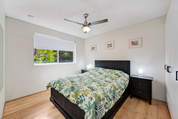 A bedroom with a bed, a ceiling fan, a window with a view of greenery, and three framed pictures on the wall.
