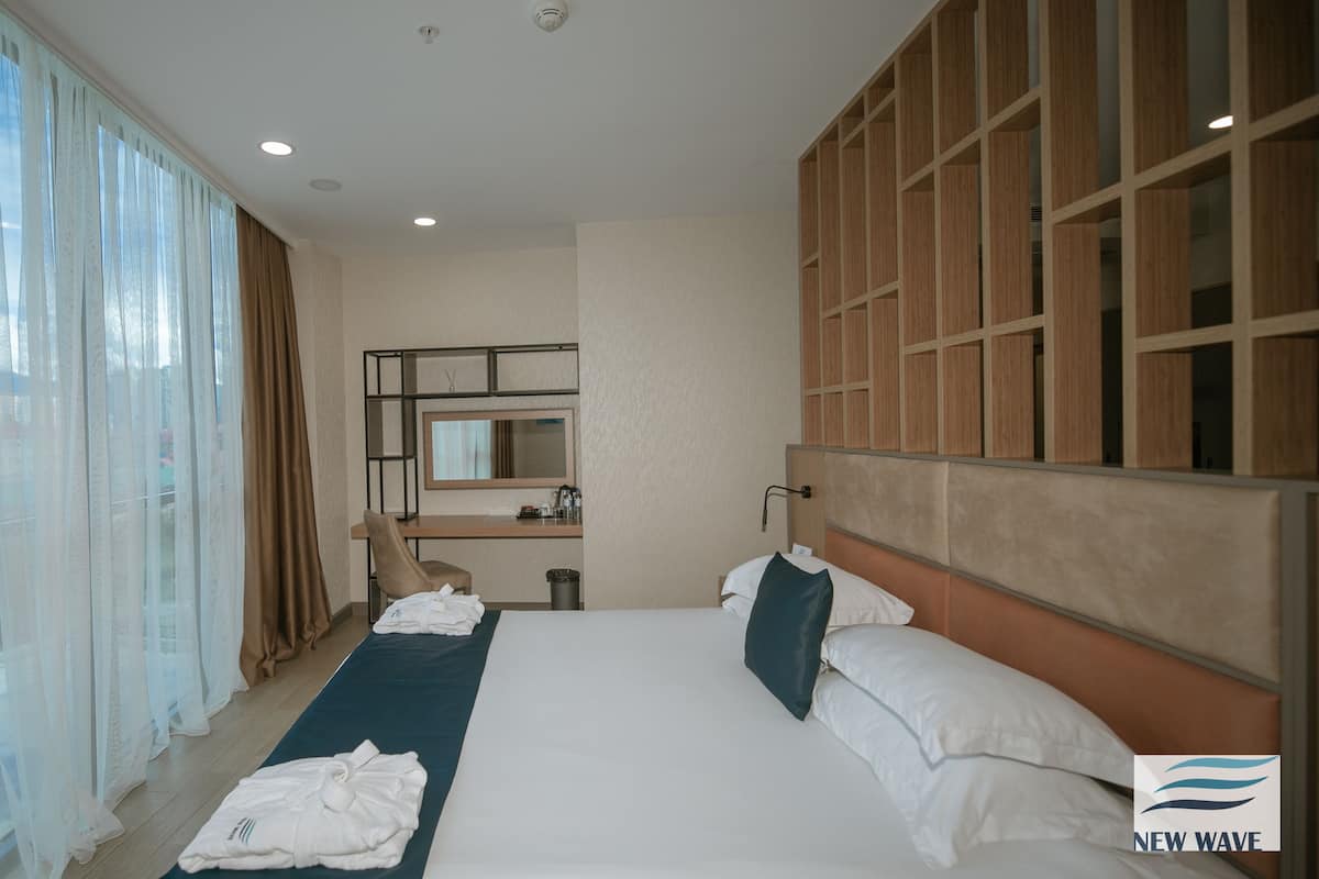 A hotel room with a large bed, a desk with a chair, a mirror, and a view of the city through sheer curtains.