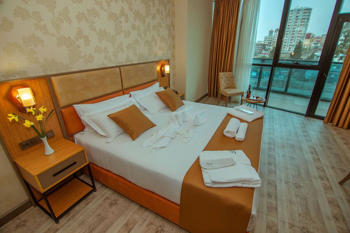 A hotel room with a large bed, a nightstand with a lamp, a chair, and a view of the city through large windows.
