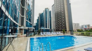A rooftop pool with lounge chairs, a pool table, and a view of modern high-rise buildings.