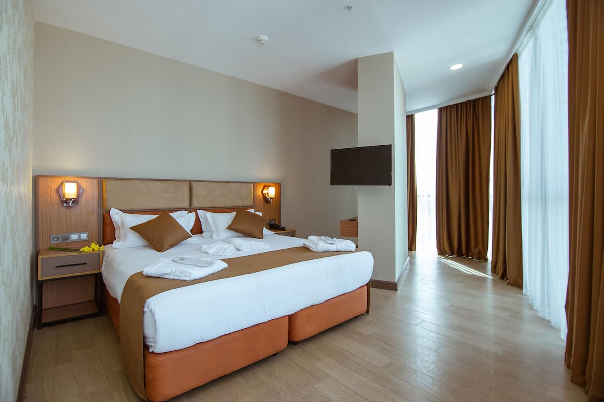 A hotel room with a large bed, two bedside tables, a flat-screen TV, and a large window with curtains.