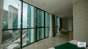 A modern hotel room with large floor-to-ceiling windows, a green bed, a chair, and a small table.