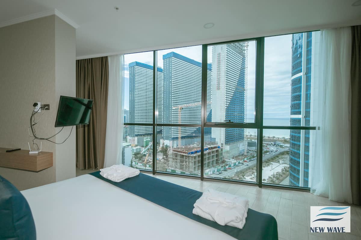 A hotel room with a large bed, a TV mounted on the wall, and a view of tall buildings through large windows.