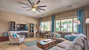 A spacious living room with a large sofa, a coffee table, and a television. There is a ceiling fan, a dining table with chairs, and a bookshelf. - Modern Azure Home w/ Beautiful Patio, Pool & Spa! (Goodyear)