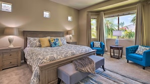A hotel room with a large bed, two bedside tables with lamps, a bench, a blue armchair, and a view of palm trees through the window. - Modern Azure Home w/ Beautiful Patio, Pool & Spa! (Goodyear)