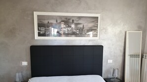 A bedroom with a large bed, a cityscape artwork on the wall, a radiator, and a nightstand with a lamp.