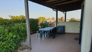 A covered patio with a table and chairs, surrounded by greenery and a view of the sea.