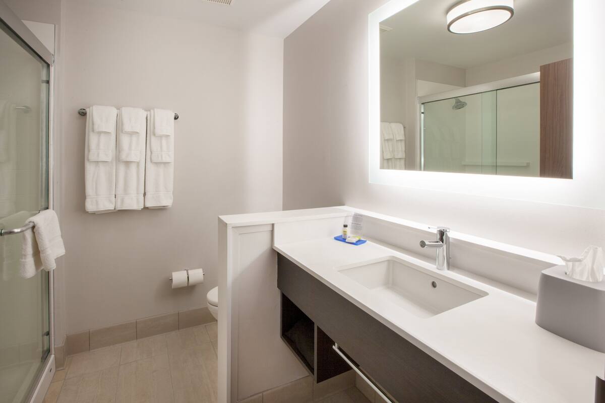 Un bany modern amb un espai de banyera de vidre, un lavabo blanc amb un mirall a dalt i una barra de tovalloles amb diverses tovalloles.