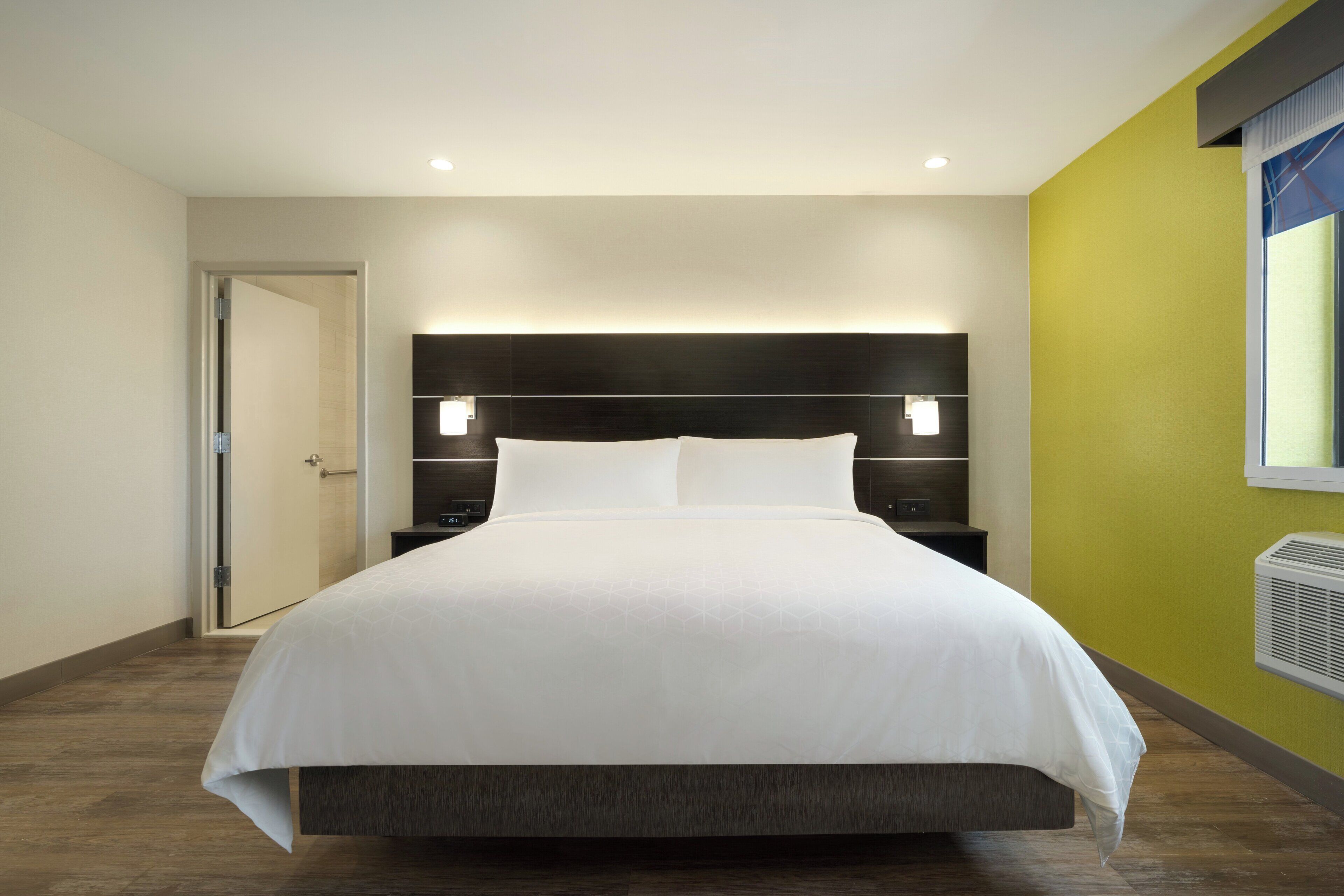 Habitación de hotel con una cama grande, una mesita de noche, una lámpara de pared y un ventanal con persianas.