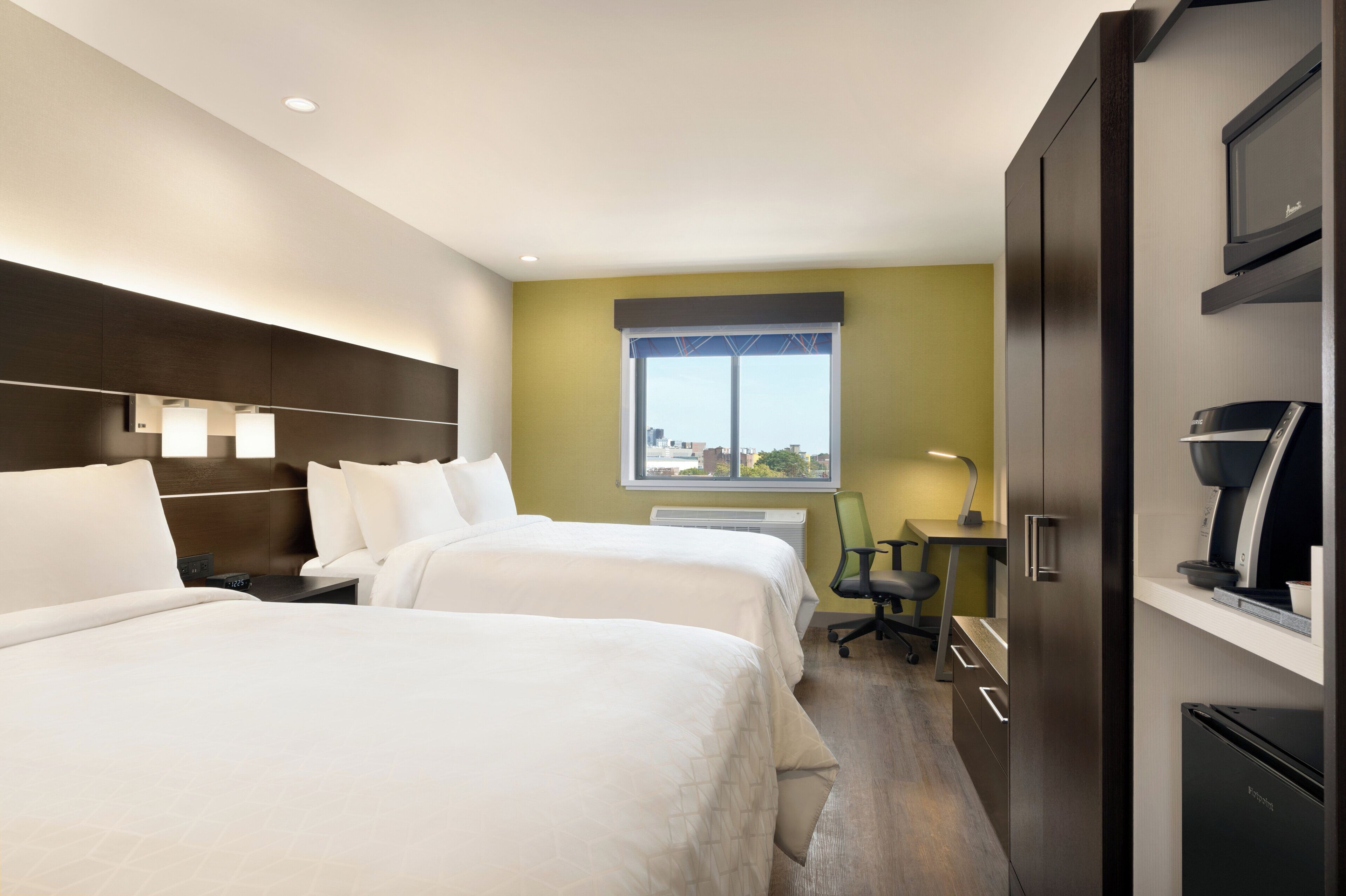 Habitación de hotel con dos camas, un escritorio, una silla, un ventanal con vista a la ciudad y un minibar.