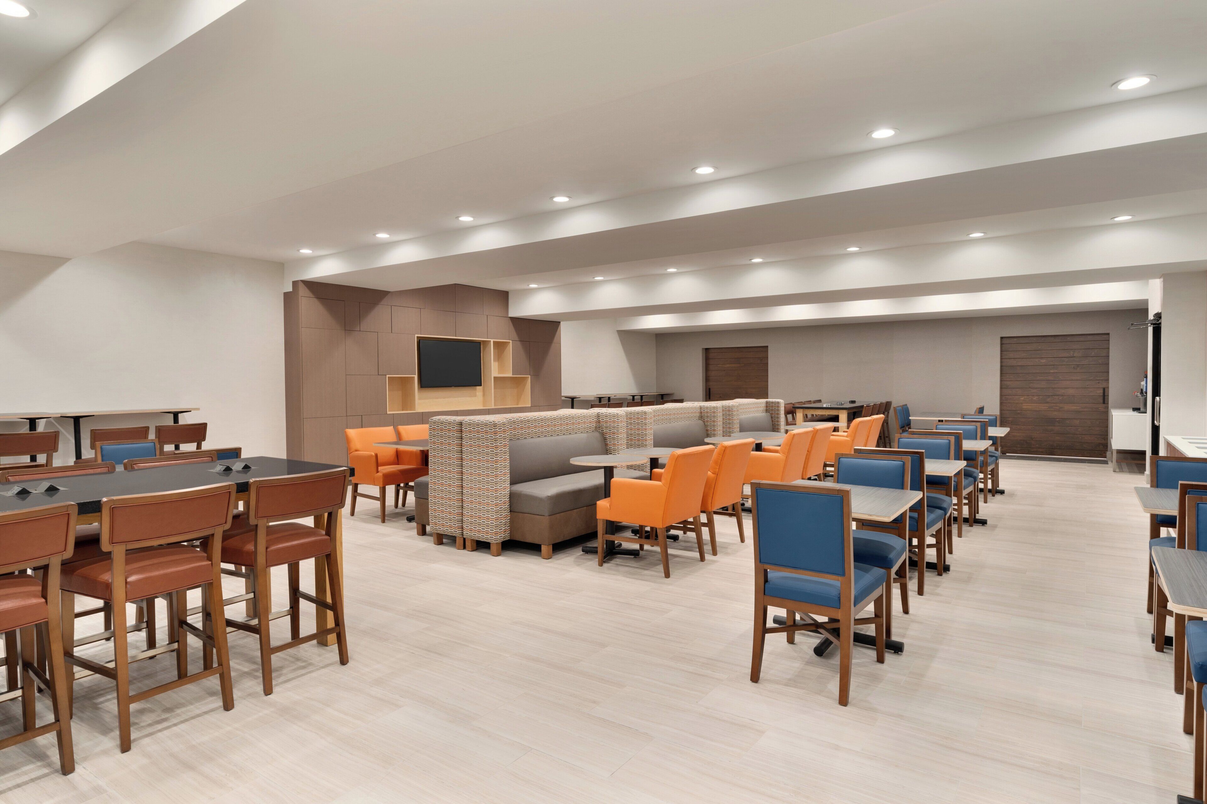 A modern hotel lobby with multiple seating areas, including tables and booths, and a flat-screen TV mounted on the wall.