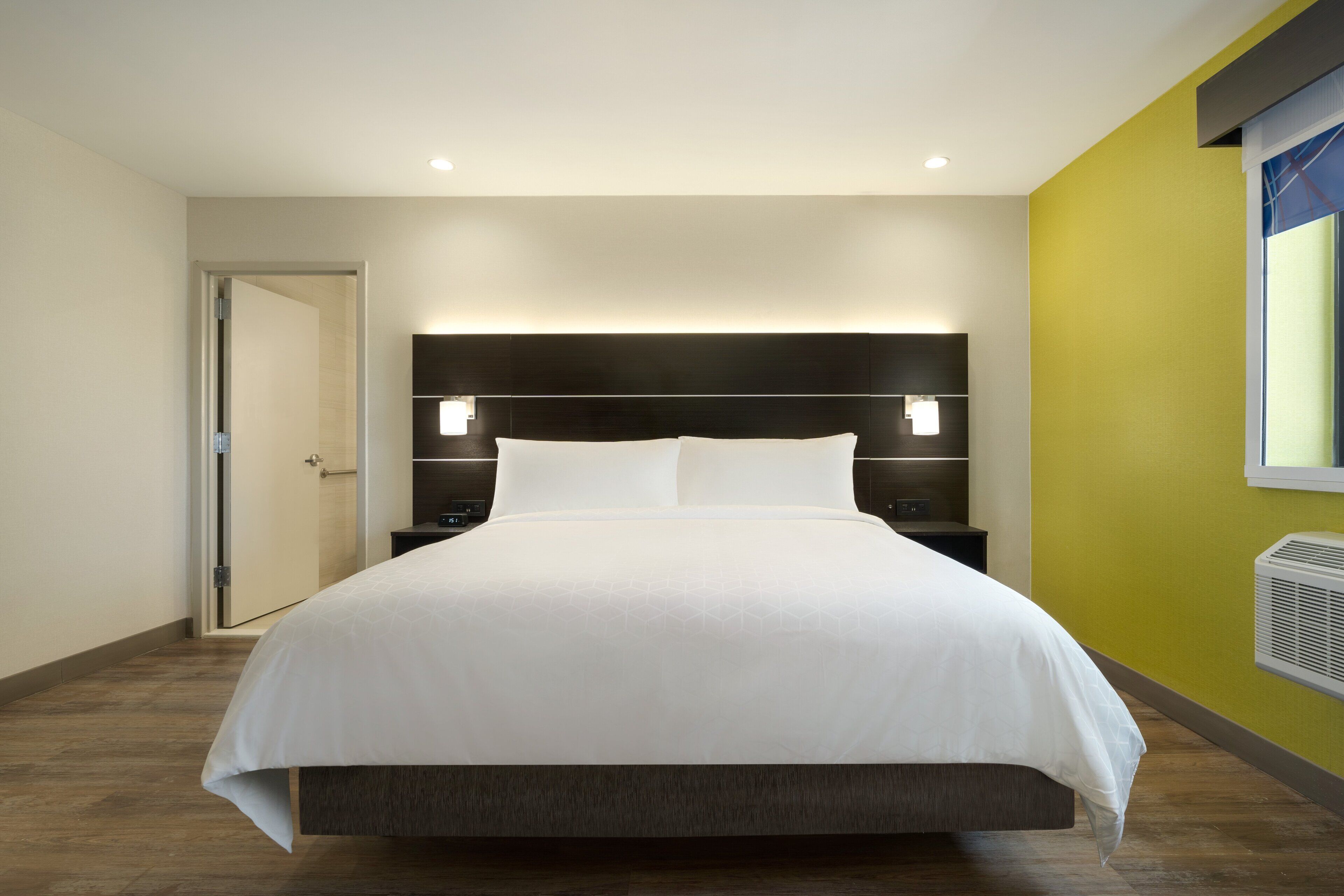 A modern hotel room with a large bed, a dark headboard, bedside tables, and a window with an air conditioning unit.