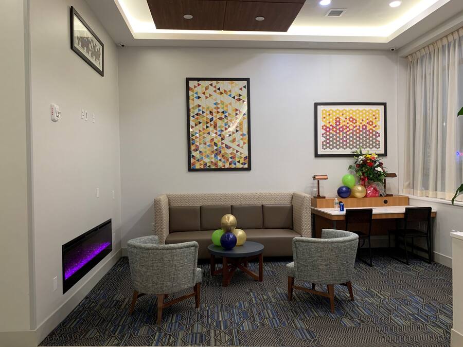 A modern hotel room with a sofa, two armchairs, a small table, and a desk with a lamp. There are two framed artworks on the walls, a fireplace, and a window with curtains.