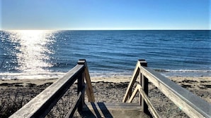 Ein Holzsteg führt zu einem Strand mit freiem Blick auf das Meer, und die Sonne spiegelt sich im Wasser.