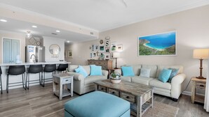 Un salon moderne comprenant un canapé, un pouf, une table basse et un coin repas avec des chaises et un comptoir de cuisine.